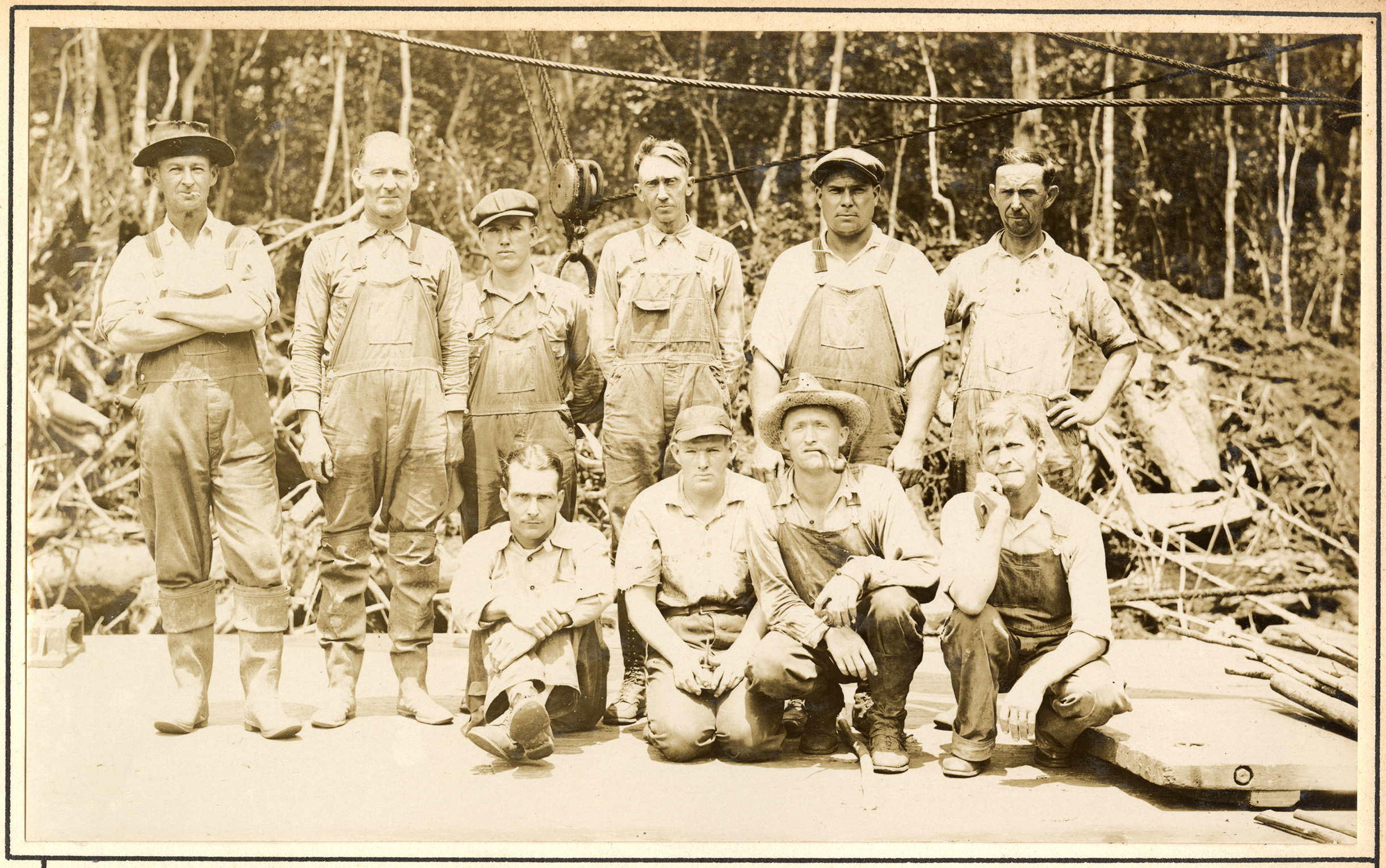 Group of 10 men wearing overalls in 2 rows in the woods